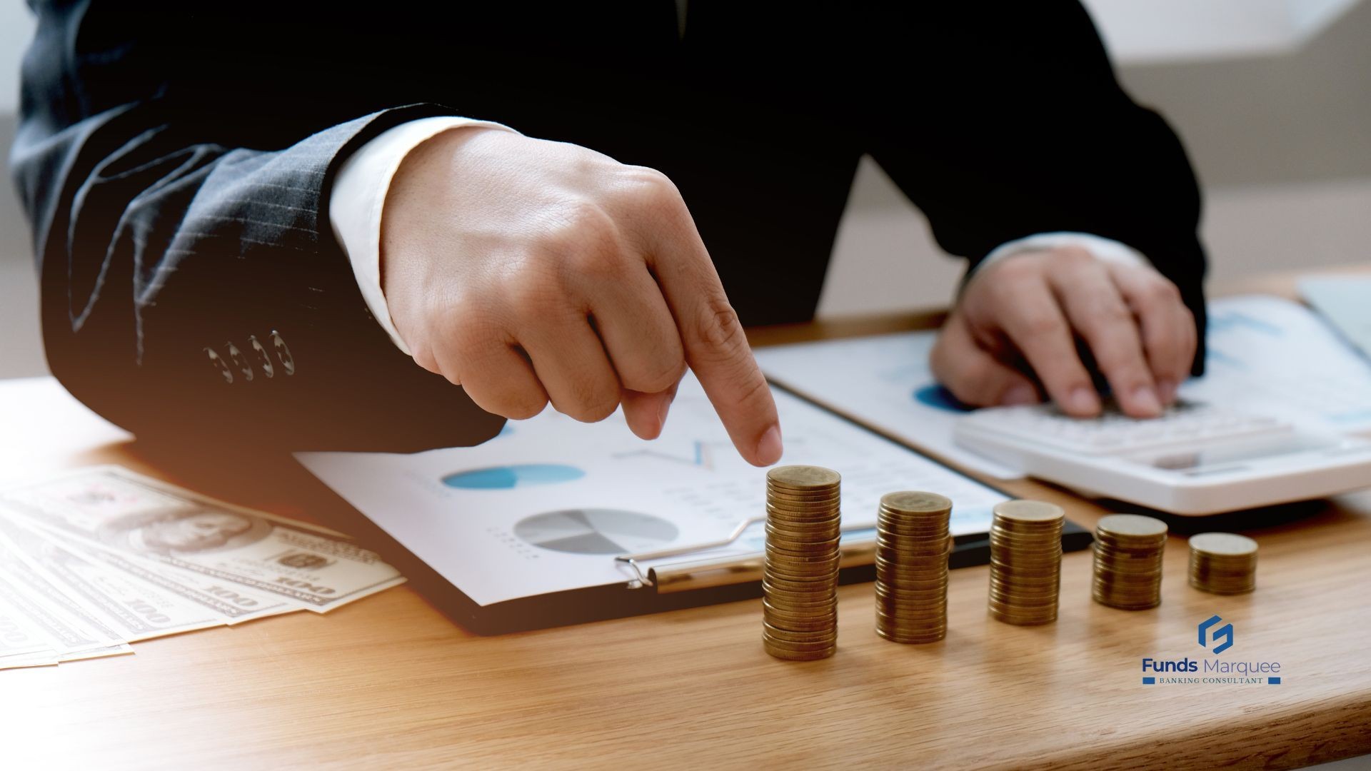 Person in a suit stacking coins on a table with financial graphs and a calculator in the background.